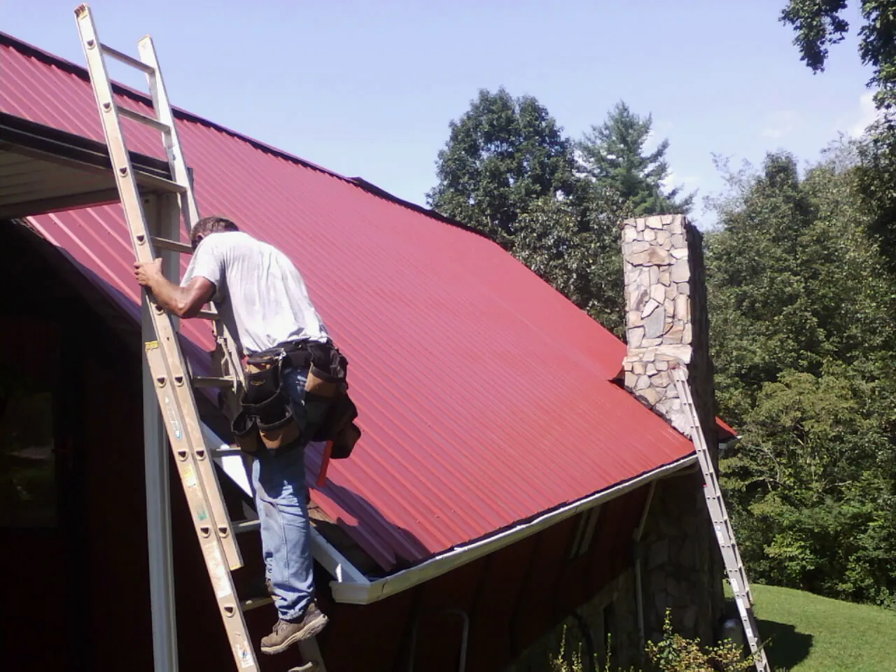 Roofer on a ladder beside a red metal roof and stone chimney — Soffit & Fascia Repair by Roofing Brothers in Big Bear Lake